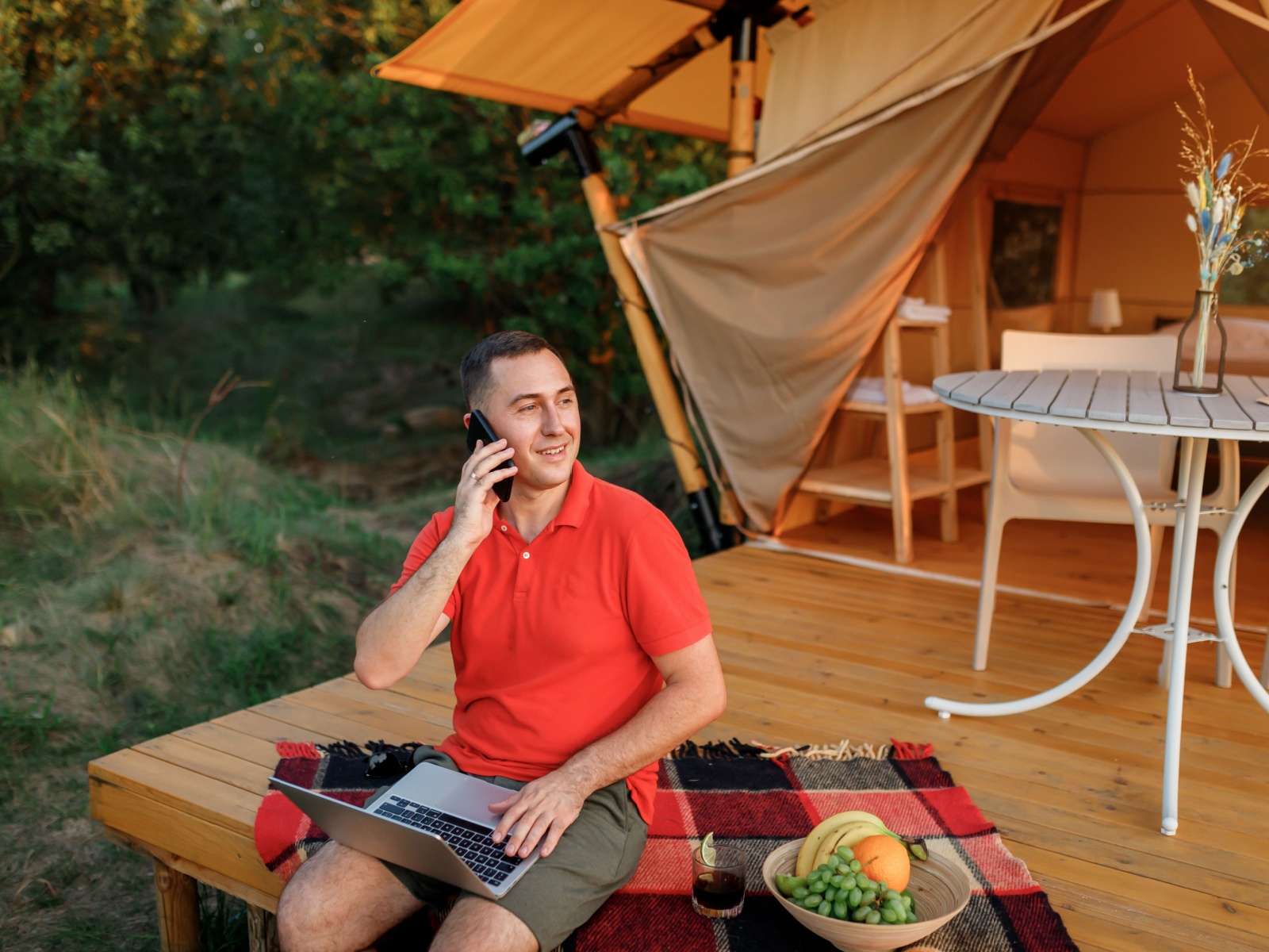 Happy businessman freelance using laptop while talking by phone on cozy glamping tent in a summer day.