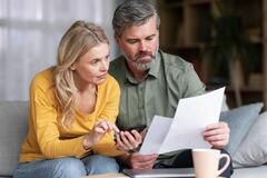 Married Middle Aged Couple Planning Budget Together, Reading Papers And Calculating Spends While Sitting On Couch In Living Room