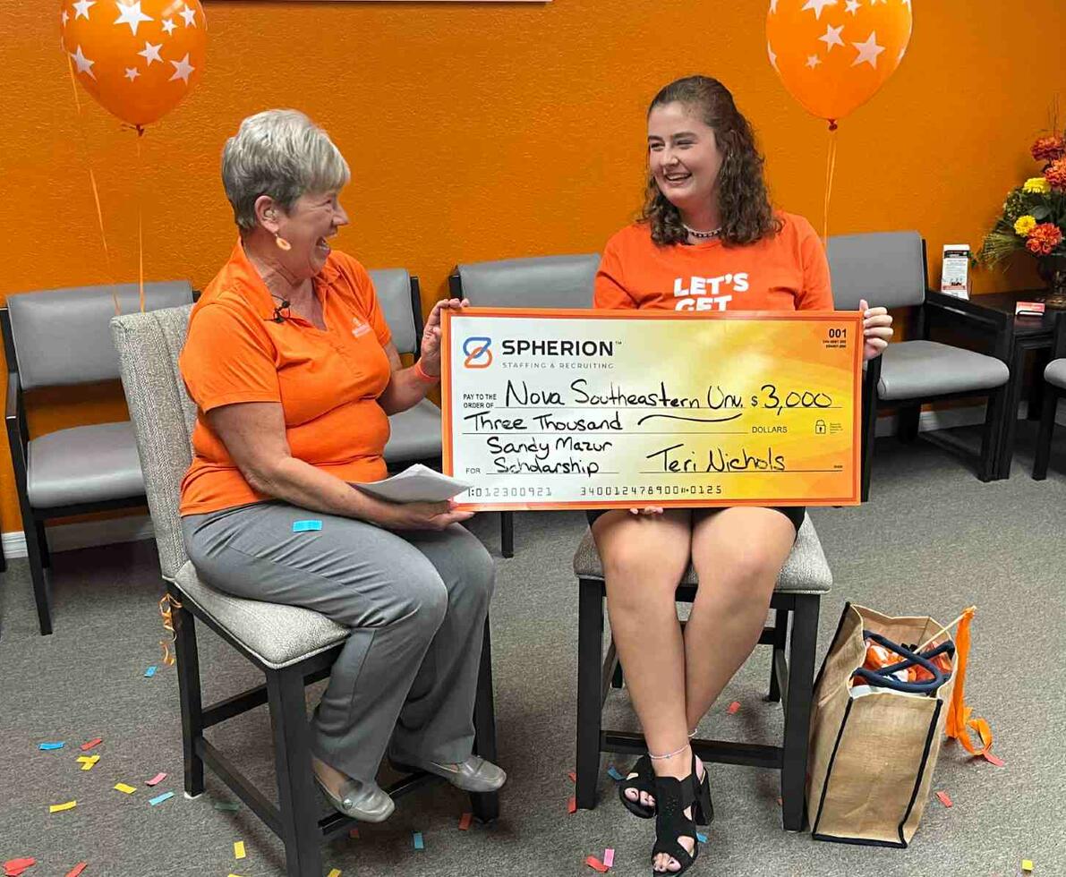 Two women in orange shirts sitting while holding a giant check